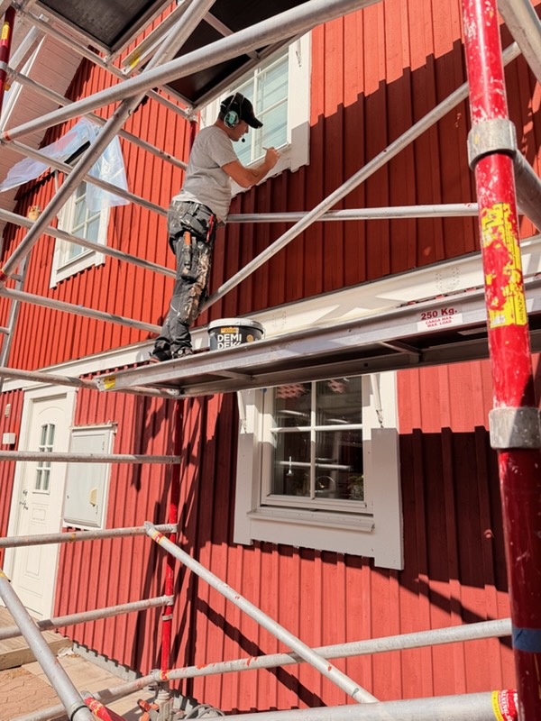 Målare arbetar från ställning längs en röd träfasad i Nässjö under pågående ommålning